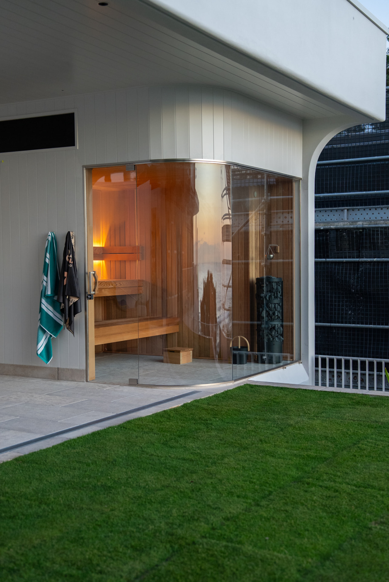 Custom curved glass sauna opening onto a lawn and covered terrace